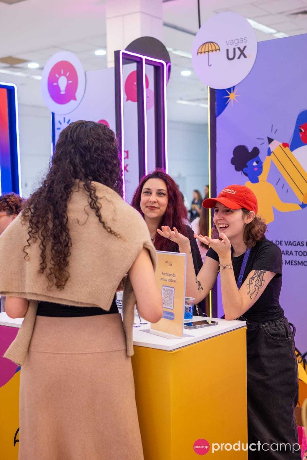 Pessoas da VagasUX conversando com uma visitante no estande da comunidade.